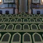 Green prayer carpet (musalla) with multiple rows of golden arch patterns in a mosque interior.